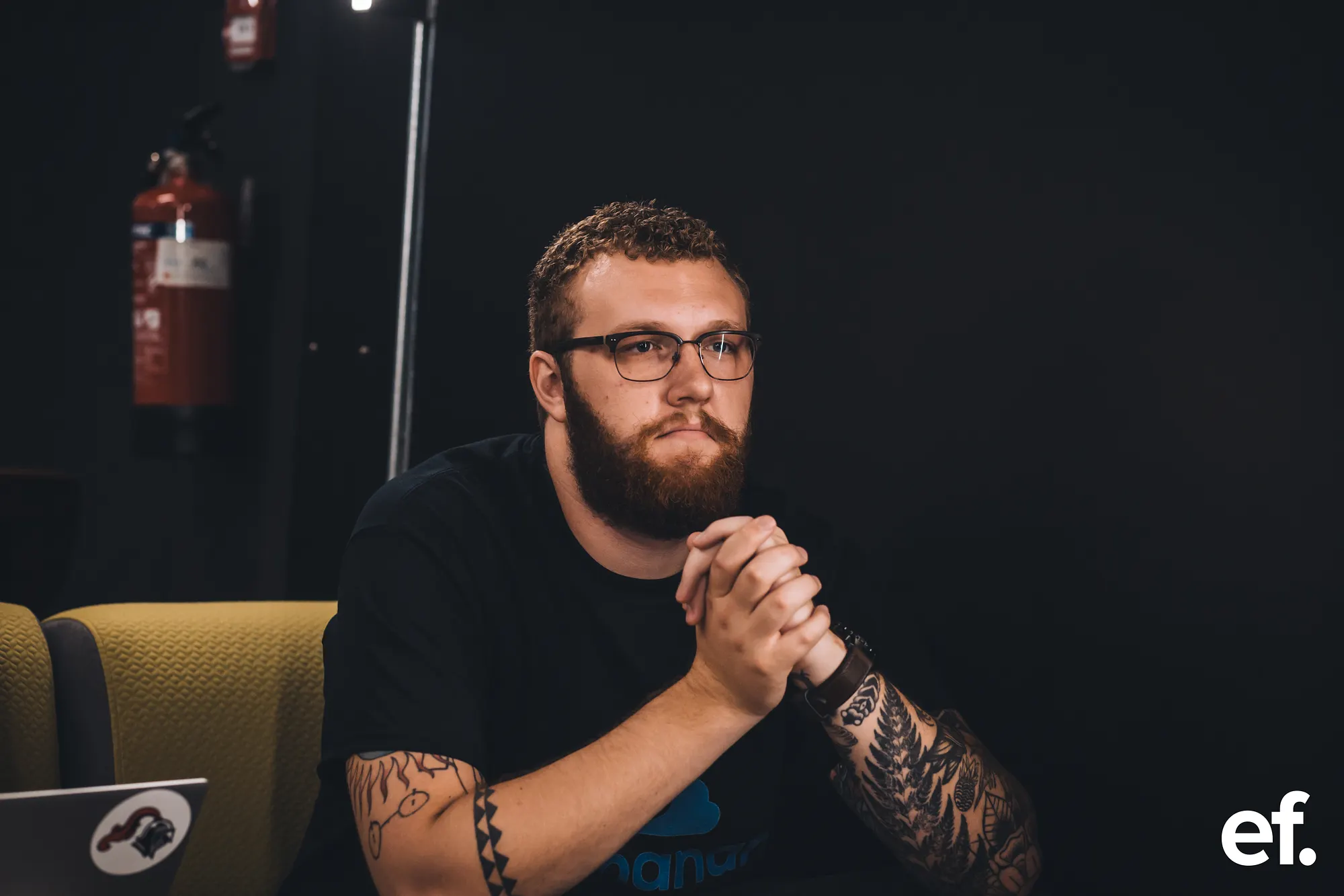 Alex Nodeland sitting in the darkened auditorium before pitching, hands clasped, wearing an Archanan t-shirt.