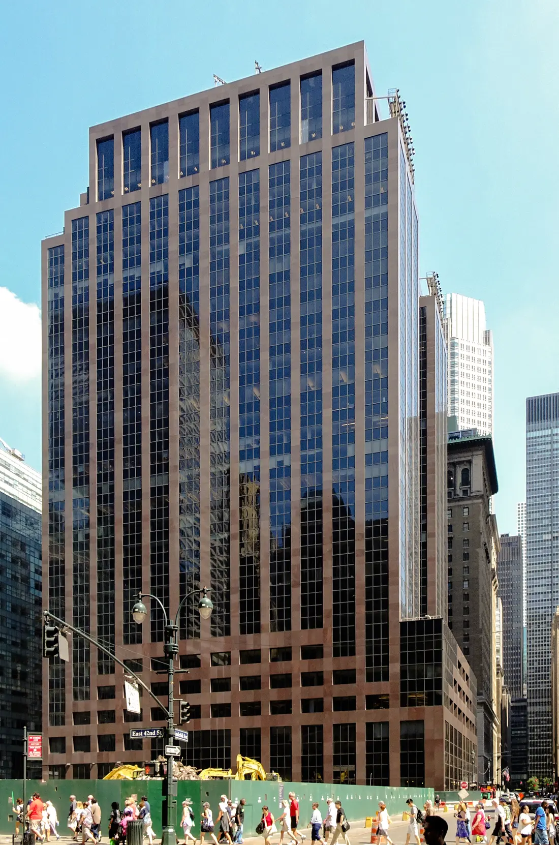 335 Madison Avenue seen from across East 42nd Street: a bronze-and-blue glass tower stepping back as it rises, with pedestrians and green construction hoarding along the sidewalk at its base.