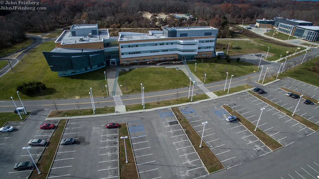 An aerial view of the Center of Excellence in Wireless and Information Technology at Stony Brook's research and development park: a low building with a curved dark-glass wing, backed by autumn woods and fronted by an almost empty parking lot.