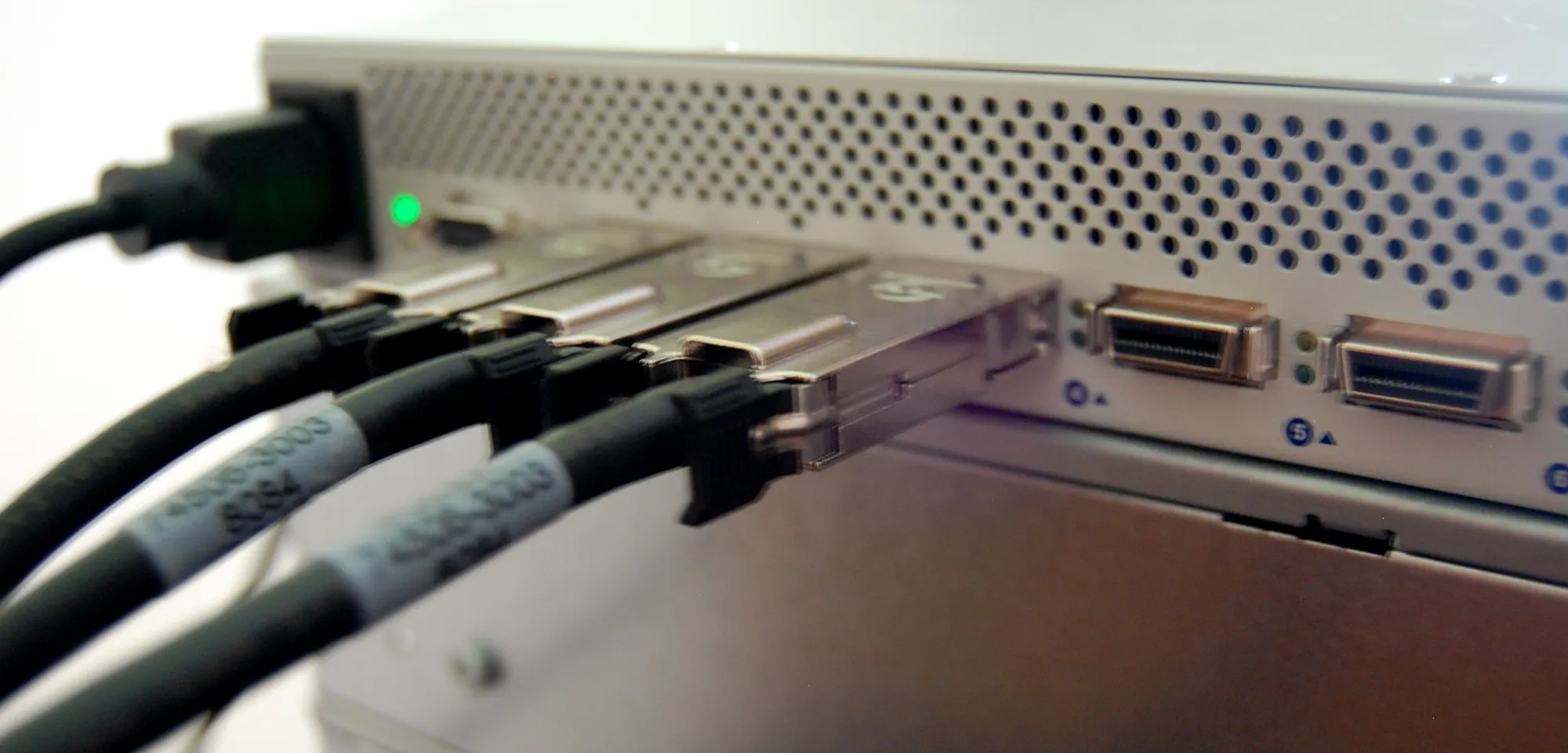Close-up of an InfiniBand switch with thick black copper cables plugged into its ports, a green status light showing on the left.